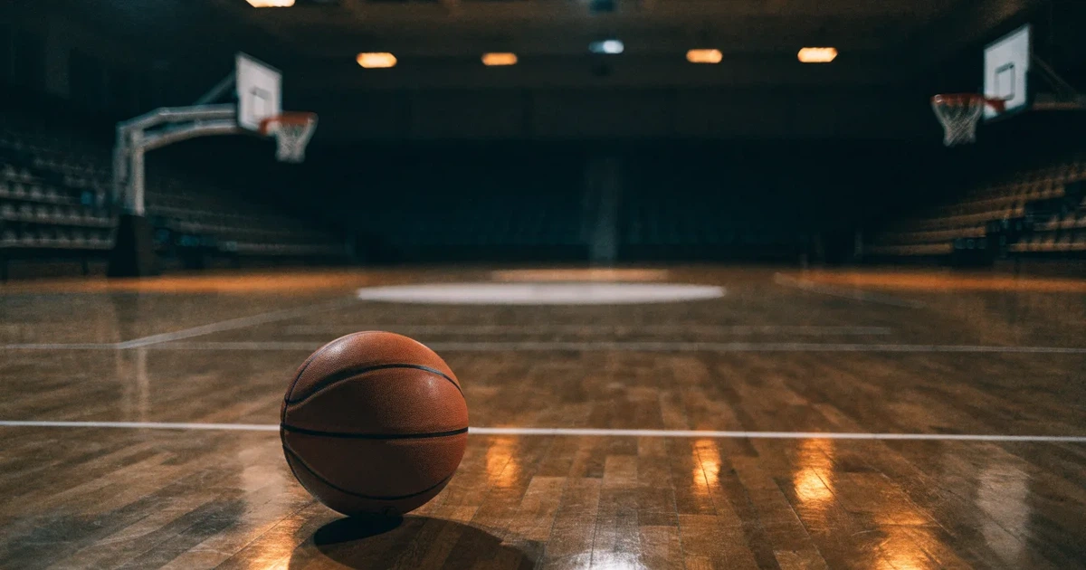 Ein orangefarbener Basketball liegt auf dem Parkett einer leeren Halle unter hellem Flutlicht, die Tribünen sind dunkel.