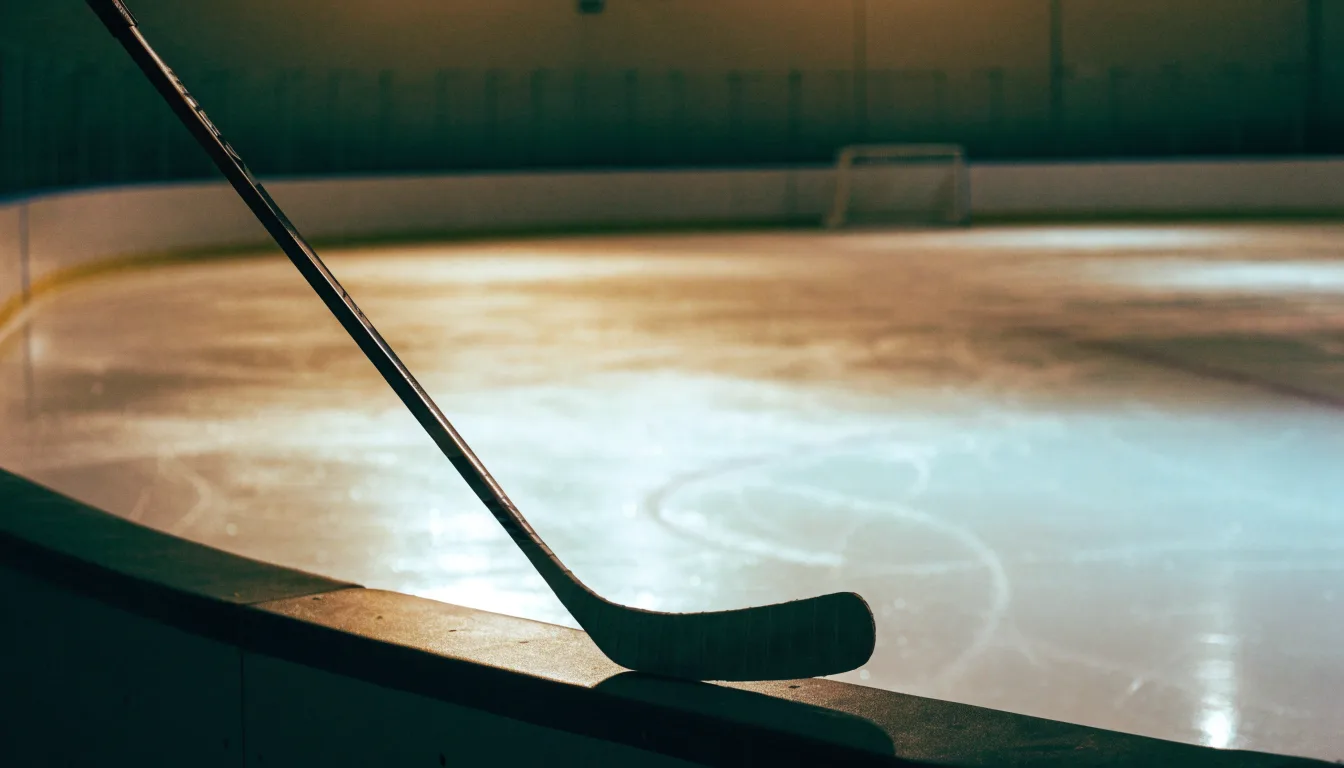 Ein Eishockeyschläger lehnt an der Bande, in der Ferne liegt die hell beleuchtete Eisfläche einer Halle unter Flutlicht.