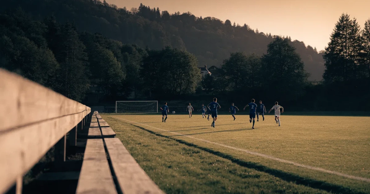 Ein kleiner Fussballplatz mit einfacher Holztribüne und Rasenstreifen am Rand einer Ortschaft, im Hintergrund ein bewaldeter Hügel im Abendlicht.