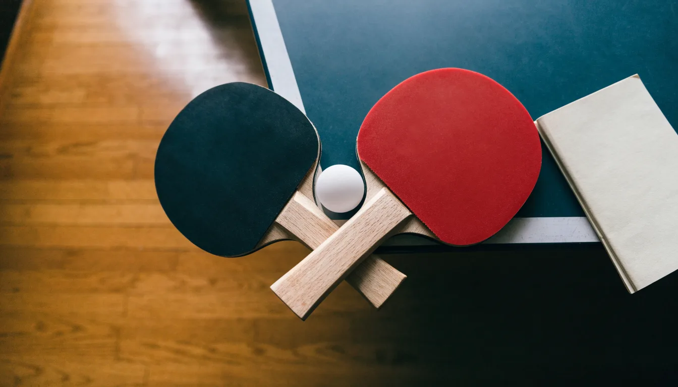 Zwei Tischtennisschläger liegen gekreuzt am Rand einer Platte, daneben ein kleiner weisser Ball.