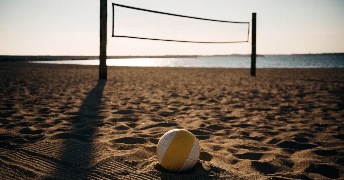 Ein Volleyball liegt im Sand eines Beachplatzes, zwischen zwei Pfosten ist ein Netz gespannt, tiefe Abendsonne wirft lange Schatten.