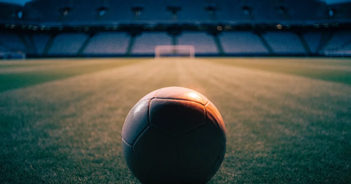 Ein Fussball liegt auf dem Anstosspunkt eines leeren Rasenplatzes, dahinter ragen die Ränge eines grossen Stadions in der Abenddämmerung auf.