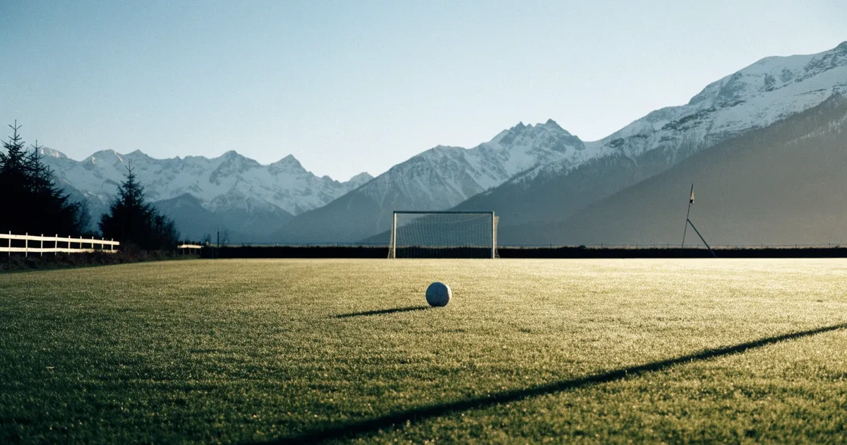 Ein Dorf-Sportplatz mit einem Ball am Strafraumrand, dahinter schneebedeckte Alpengipfel im klaren Morgenlicht