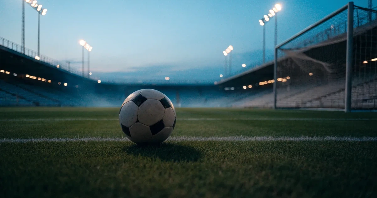 Ein Fussball liegt auf dem Anstosspunkt eines leeren Rasenplatzes in der Abenddämmerung, im Hintergrund leuchten Flutlichtmasten.