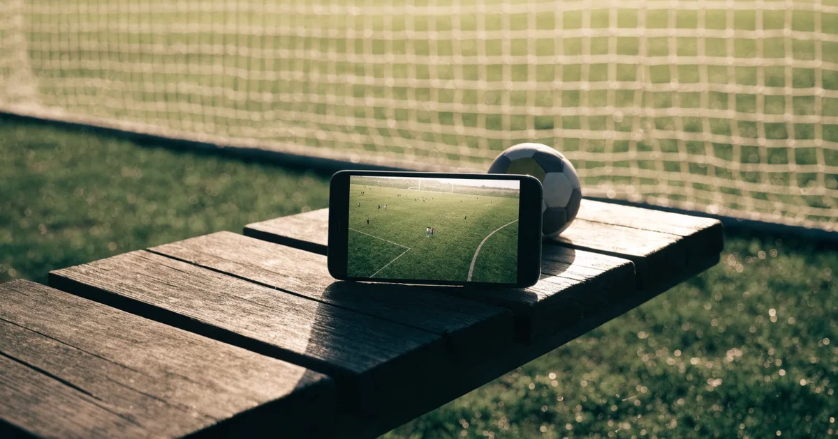 Ein Smartphone mit dunklem Bildschirm liegt neben einem Fussball auf einer Holzbank am Rand eines Trainingsplatzes im Morgenlicht.